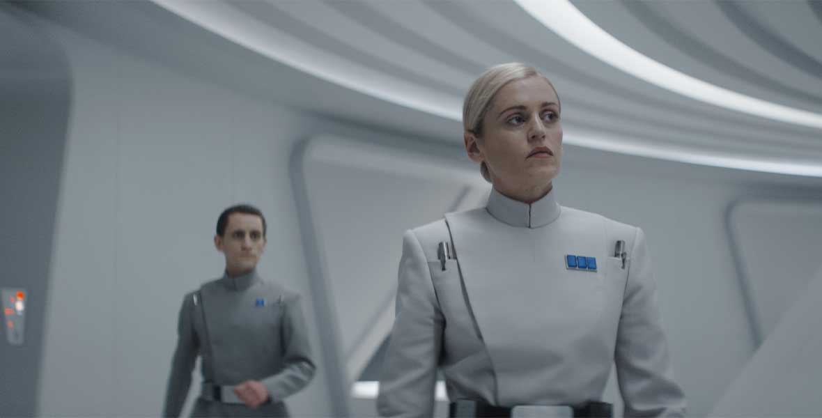 Una donna con capelli biondi in uniforme bianca. Alle sue spalle un uomo in uniforme grigia.